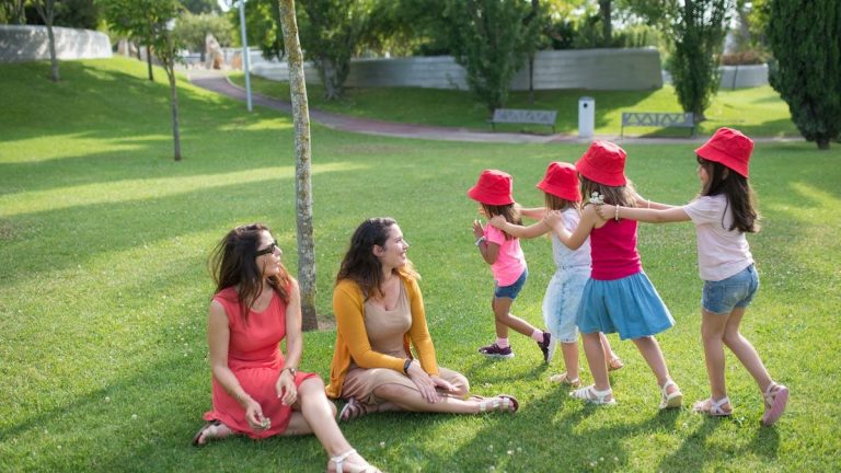 Happy children playing in a sunny park with two mothers sitting nearby, enjoying the outdoors.