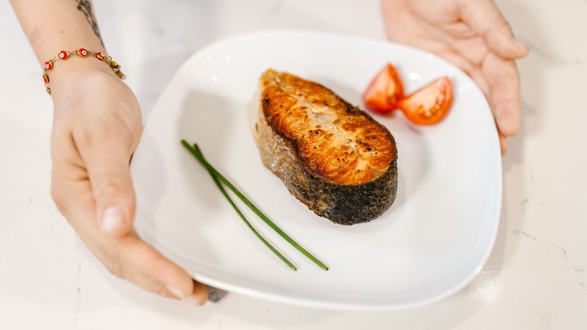 Close-up of grilled salmon served with tomatoes and chives on a plate, held by tattooed hands.